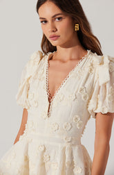A woman is wearing an elegant cream-colored dress with intricate floral embroidery. The dress features a deep V neckline with a scalloped trim, short puffed sleeves, and a fitted waist. The fabric has a textured, embroidered floral pattern throughout. She accessorizes with bold, geometric gold earrings.